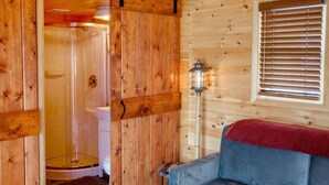 A wooden cabin interior with a sliding door leading to a shower, a white toilet, a blue sofa, and a window with blinds. - Gorgeous 2-bedroom cottage in charming Hammondsport awaits you (Hammondsport)