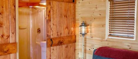 A wooden cabin interior with a sliding door leading to a shower, a white toilet, a blue sofa, and a window with blinds.