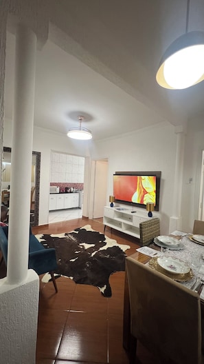 Sala de estar com televisão de tela plana, área de jantar com mesa posta para dois e cozinha ao fundo.