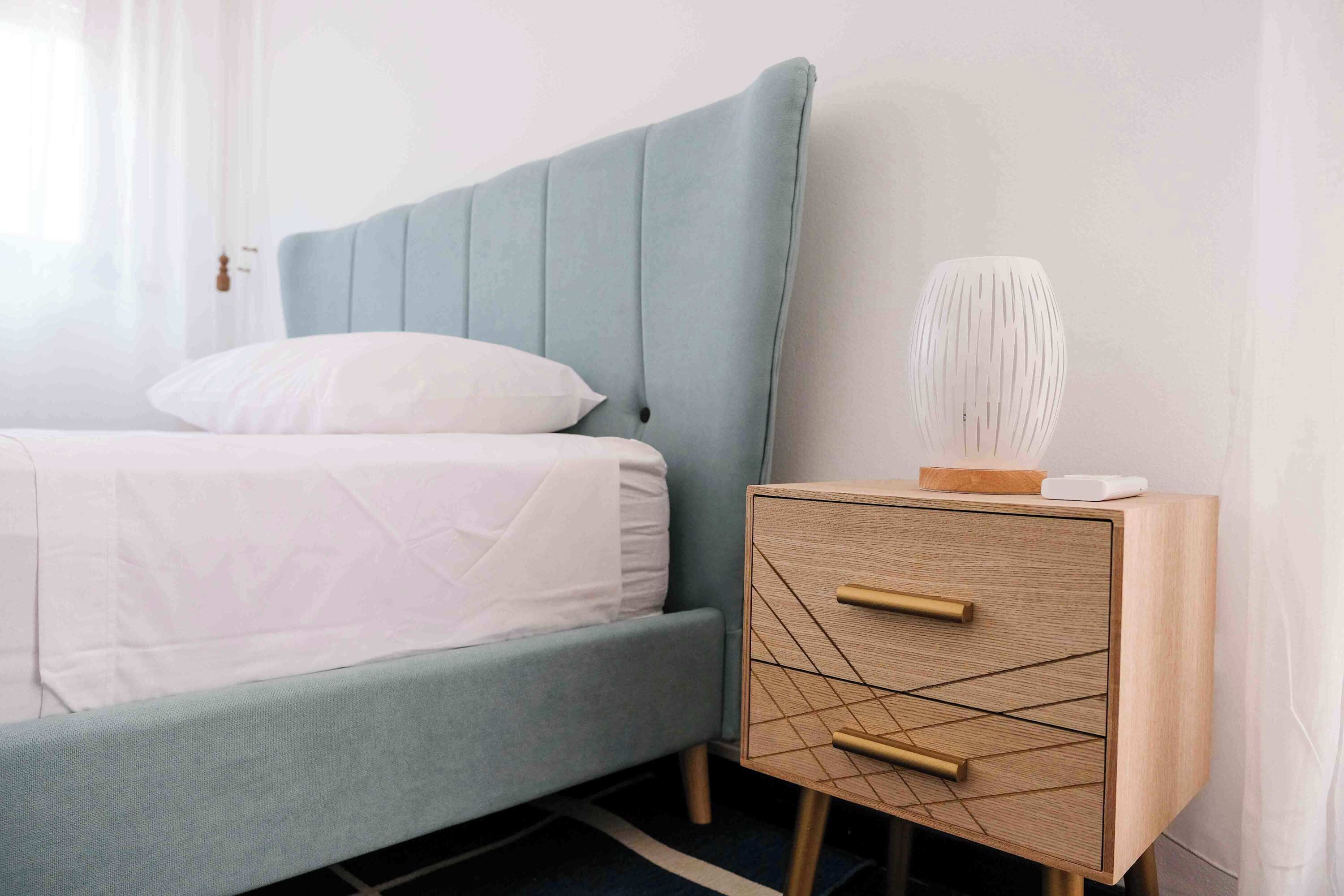 A bed with a light blue headboard, a bedside table with a geometric pattern, and a white vase on the table.
