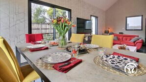 Un comedor con una mesa larga dispuesta para cuatro personas, rodeado de sillas rojas y amarillas. En la mesa hay un jarrón con flores y un televisor al fondo.
