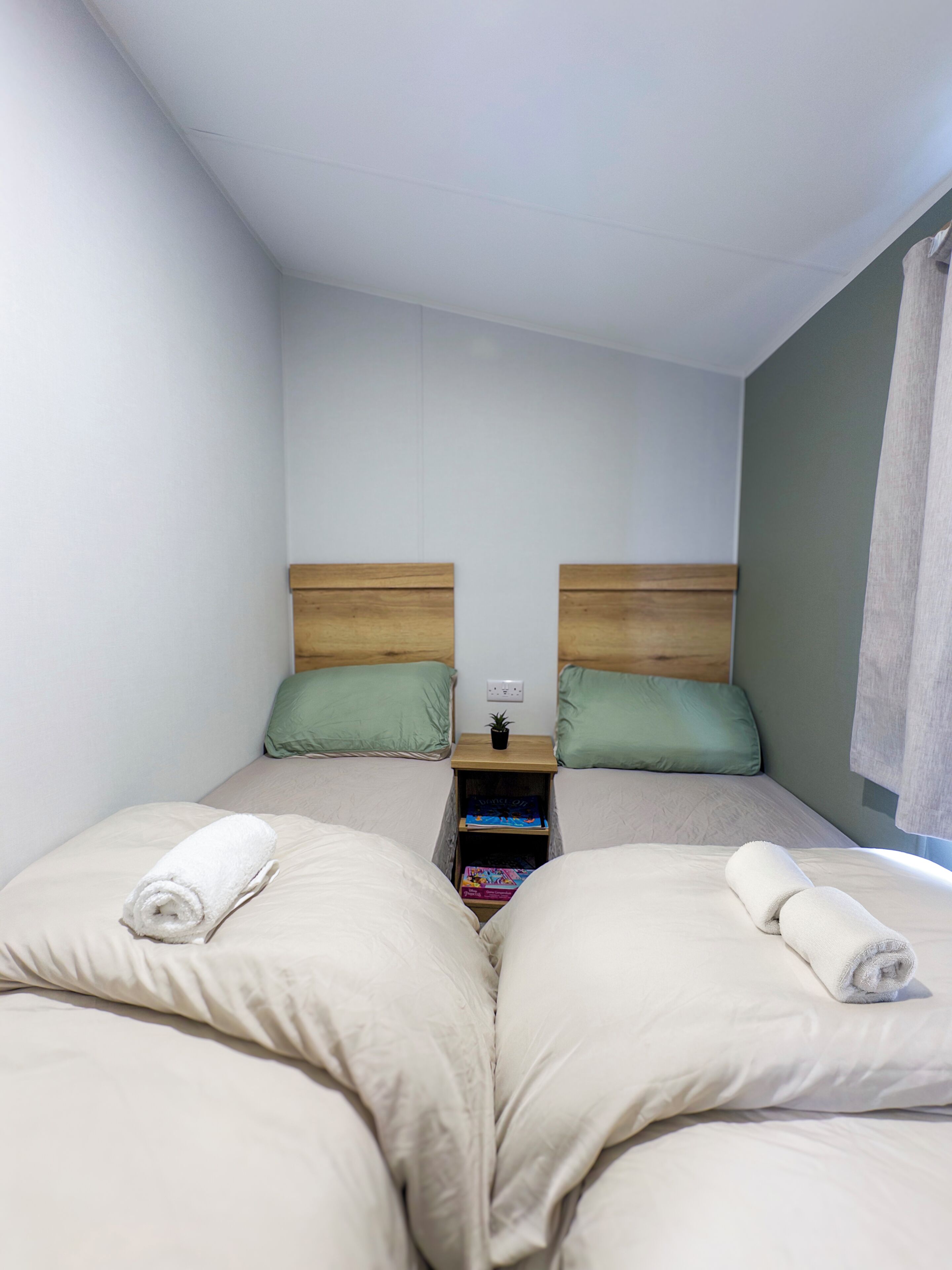 A twin room with two beds, each with a wooden headboard, green pillows, and rolled white towels. A small wooden nightstand with a plant and books is between the beds.