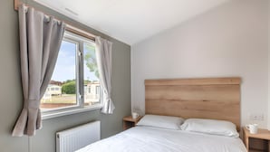 Une chambre avec un grand lit, une tête de lit en bois, une table de chevet, une fenêtre avec des rideaux et une vue sur les maisons à l’extérieur.