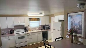 A compact kitchen with white cabinets, a stove, a microwave, and a refrigerator. There is a dining table with chairs and a vase with flowers. - Poseidon's Corner #2 of 3 units on this property (Sandusky)