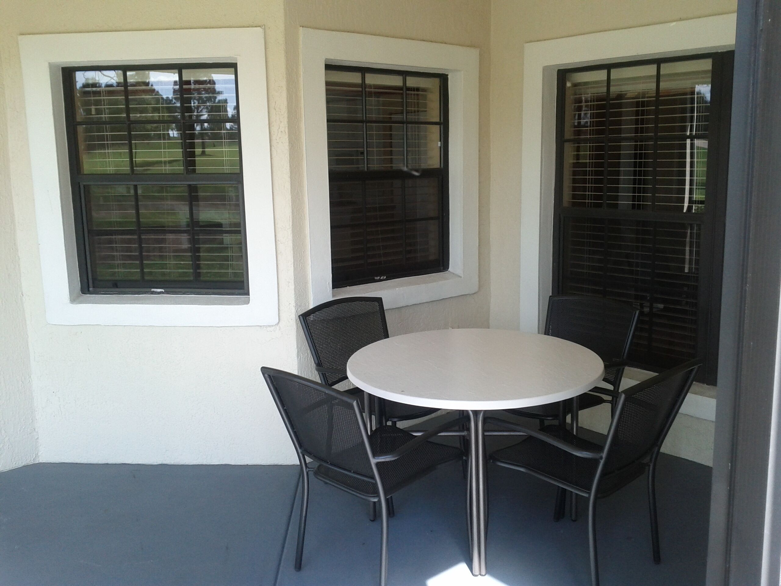 A patio with a round table and four chairs, three windows with blinds, and a concrete floor.
