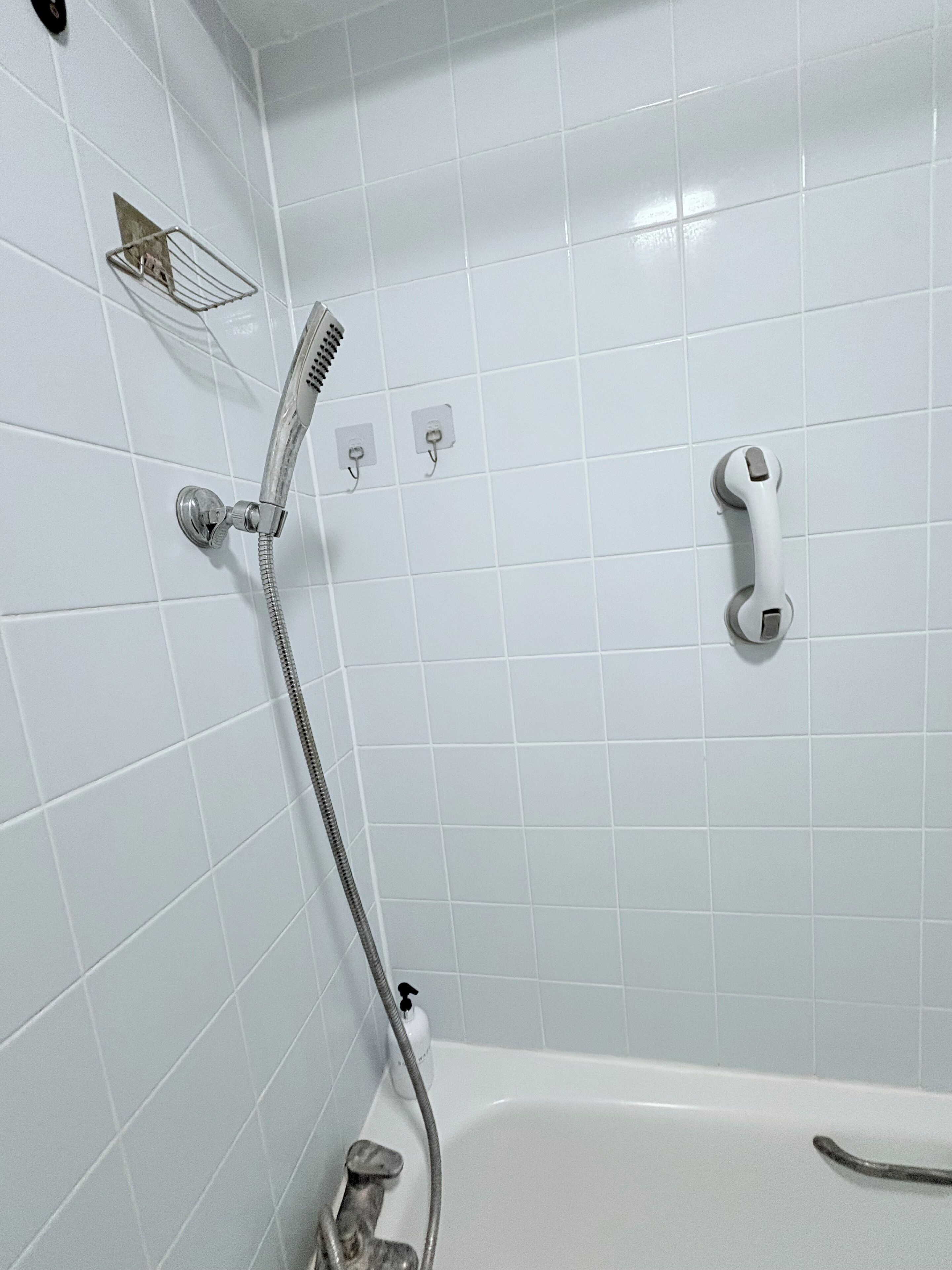A bathroom with white tiled walls, a shower with a handheld showerhead, a wall-mounted faucet, and a bathtub with a grab bar.
