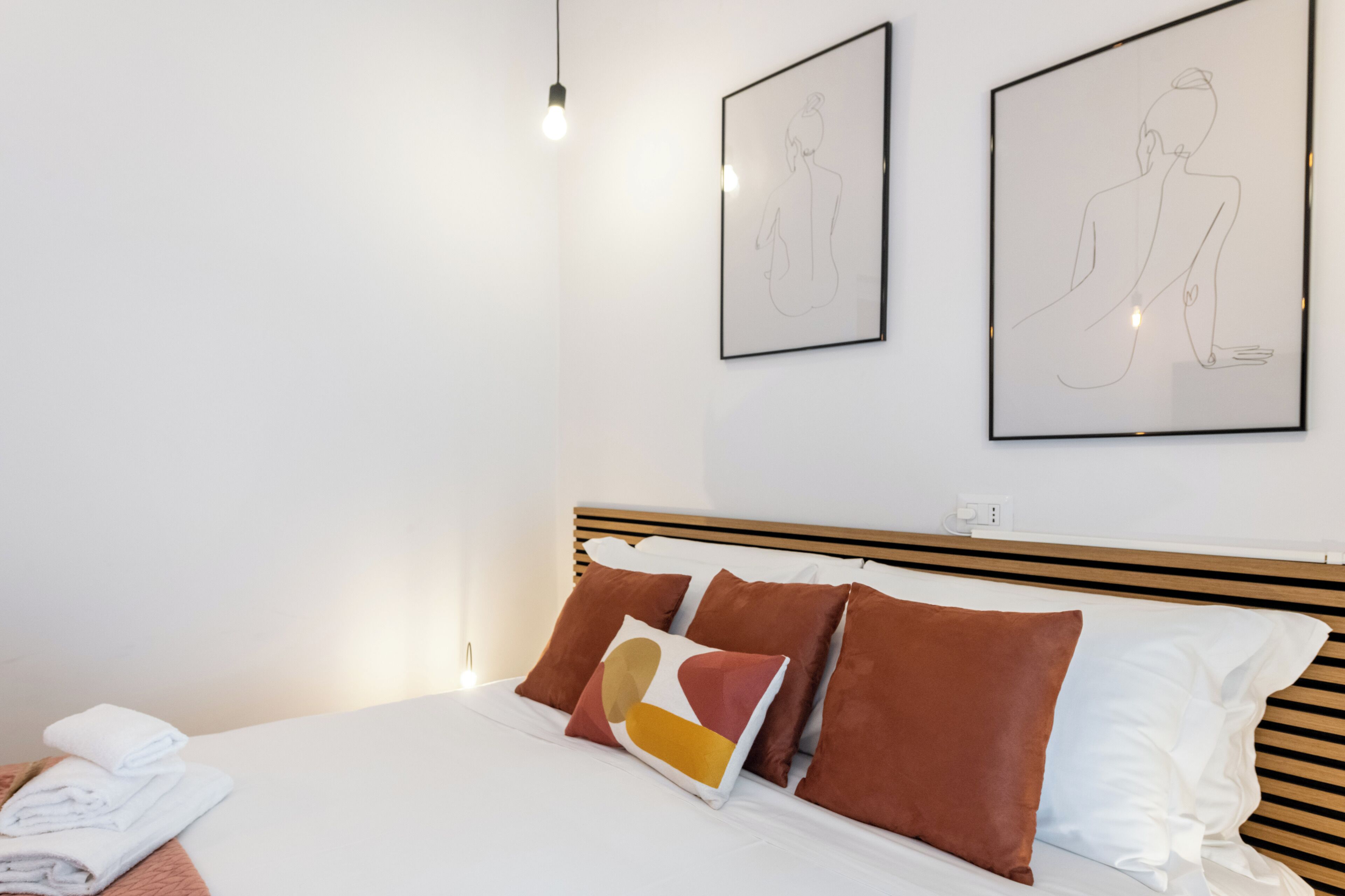 Ein ordentlich bezogenes Bett mit braunen und weißen Kissen, ein gefaltetes Handtuch und ein gerahmtes abstraktes Kunstwerk an der Wand.