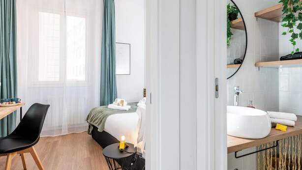 Een moderne hotelkamer met een bed, een bureau met stoel, een ronde tafel met lamp, een badkamer met wastafel en spiegel, en uitzicht op gebouwen door dunne gordijnen.