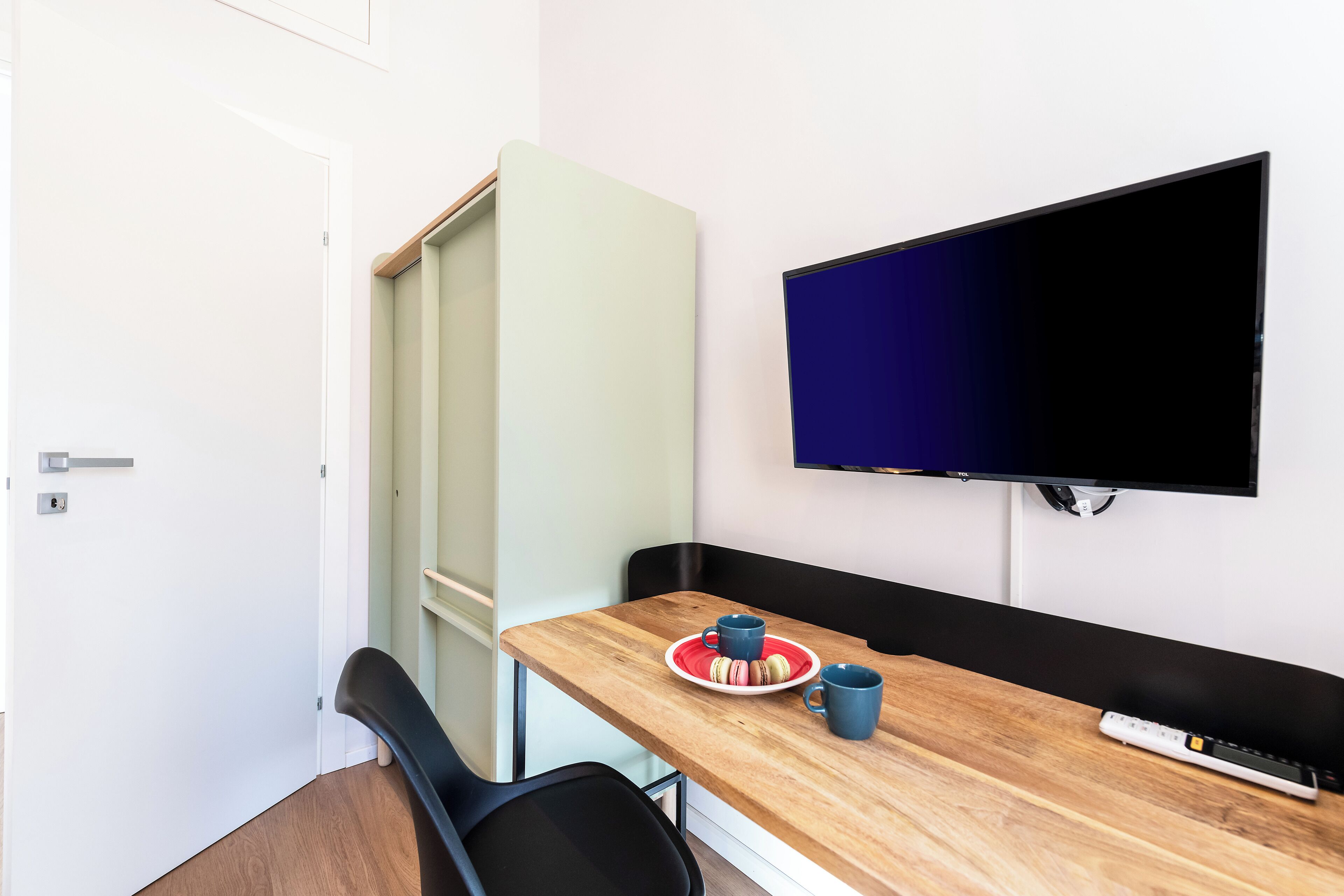 A small room with a wooden table, a black chair, a television mounted on the wall, and a door.