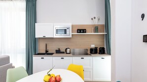 Een moderne keuken met witte kastjes, een magnetron, een spoelbak en een koffiezetapparaat. Er staat een eettafel met een bord fruit en een gele stoel.