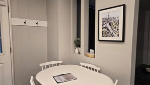 A white dining table with four chairs, a framed cityscape painting, a small vase with a plant, and a tissue box on the wall. - ​Mainstreet Hideaway | Cozy Downtown Gem in the Heart of Powassan (Powassan)