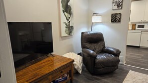 A living room with a flat-screen TV on a wooden stand, a leather armchair, a white throw blanket, and a small table with board games. - ​Mainstreet Hideaway | Cozy Downtown Gem in the Heart of Powassan (Powassan)