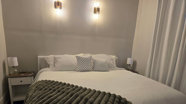 A hotel room with a large bed, two bedside lamps, a nightstand, and a window with curtains. - ​Mainstreet Hideaway | Cozy Downtown Gem in the Heart of Powassan (Powassan)
