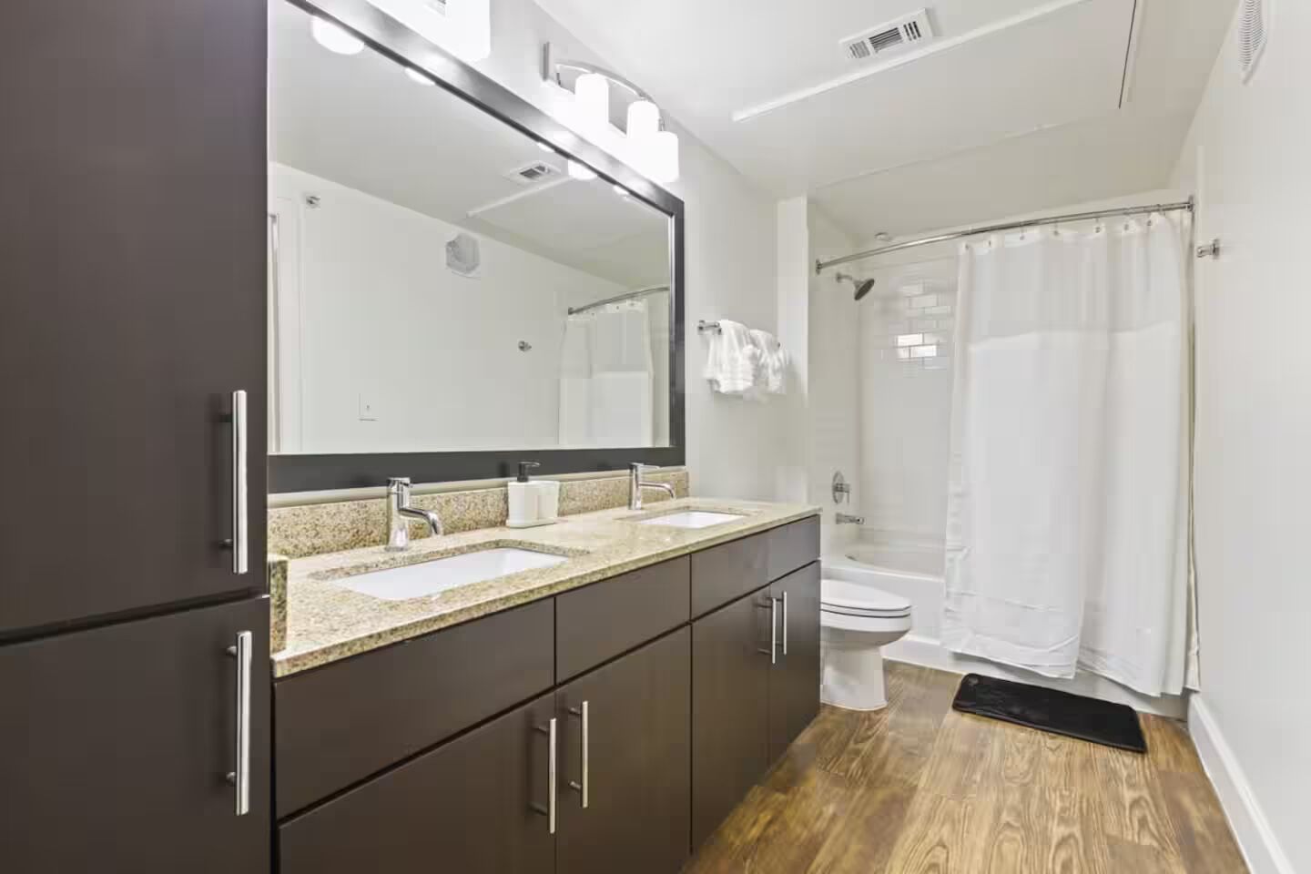 Un baño moderno con muebles oscuros, un espejo grande, dos lavamanos y una zona de ducha.