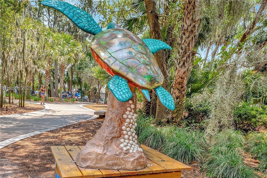 A large turtle sculpture with a textured shell and turquoise flippers, set on a wooden platform in a park with trees and a pathway.
