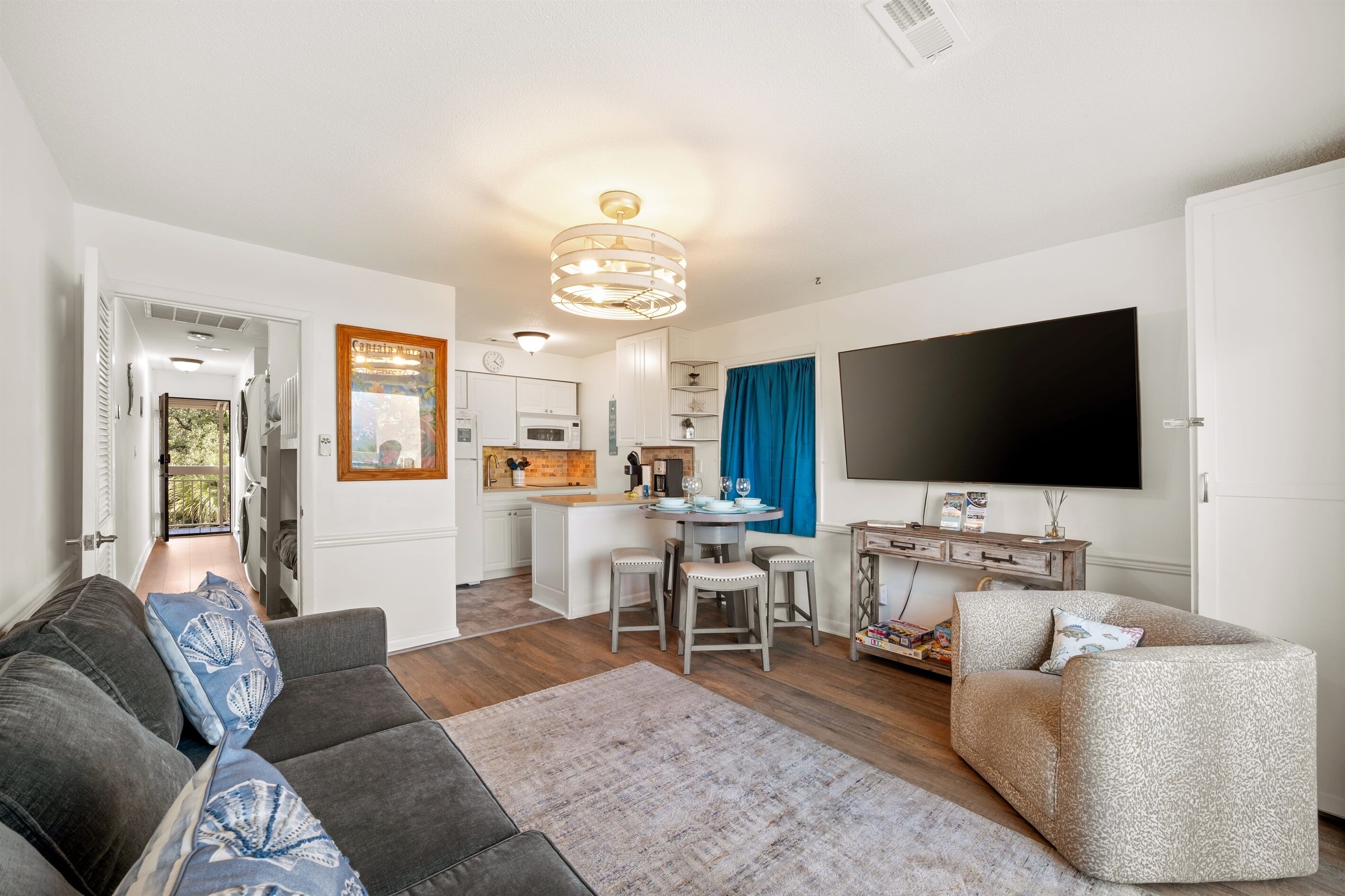 A living room with a sofa, a coffee table, and a TV. There is a kitchen in the background with a blue curtain.