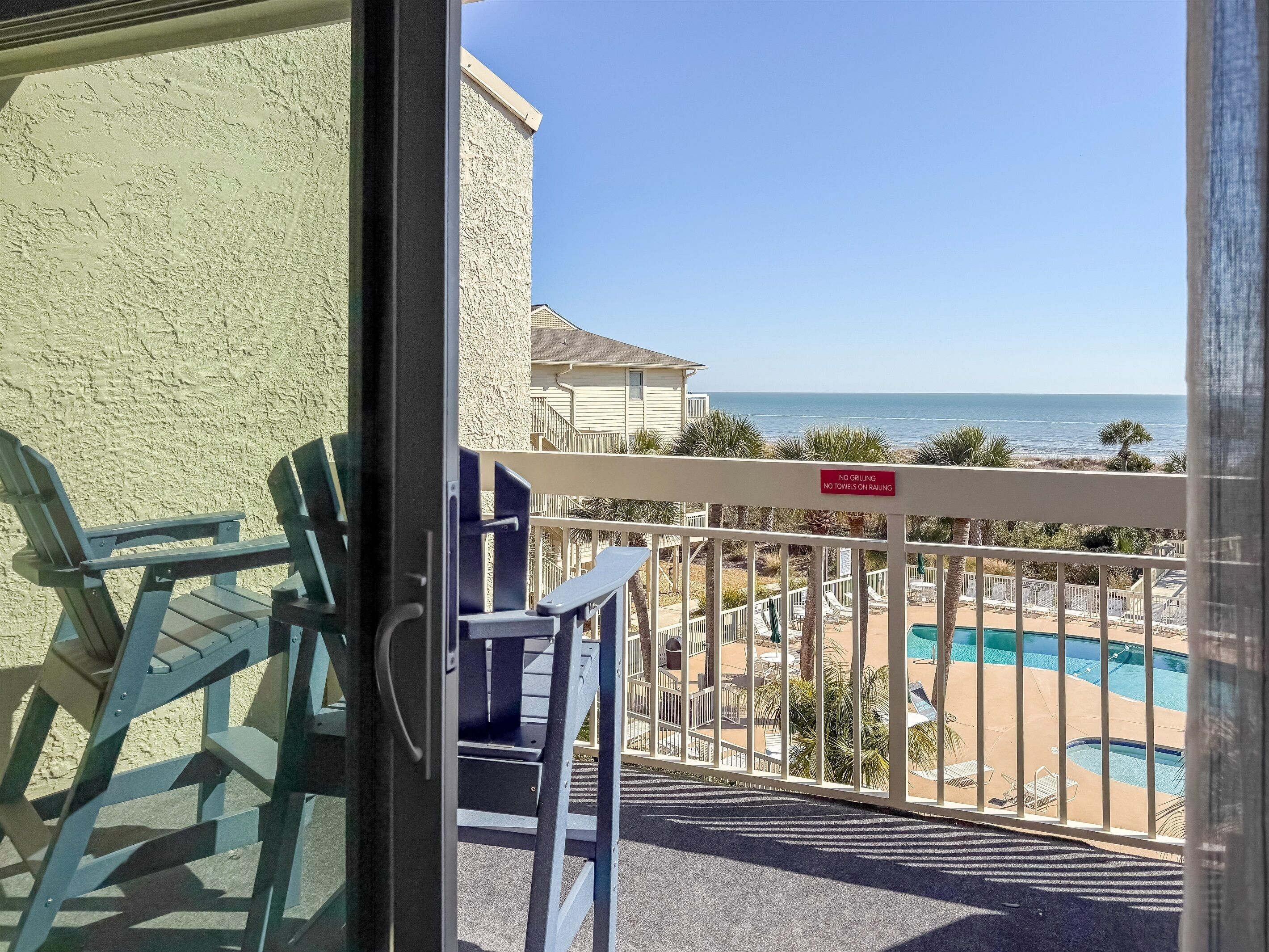 A balcony with a view of a pool and beach, a 'No Grilling' sign, and a building with a textured wall.
