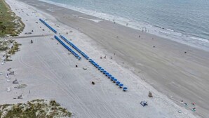 An aerial view of a beach with a long row of blue umbrellas, a sandy shore, and a calm sea. - Breakers 324 Oceanfront Hilton Head Condo Direct Beach Access (Hilton Head Island)