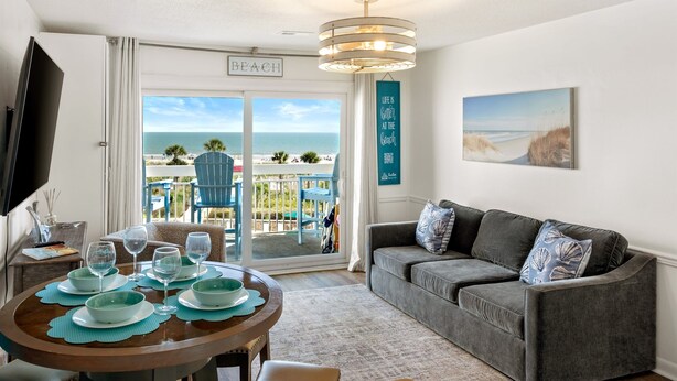 A room with a dining table set for four, a grey sofa, a TV, and a balcony with a view of the beach. - Breakers 324 Oceanfront Hilton Head Condo Direct Beach Access (Hilton Head Island)