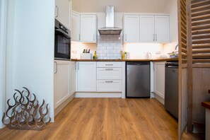 Una cocina con armarios blancos, extractor de acero inoxidable, lavavajillas y suelo de madera.