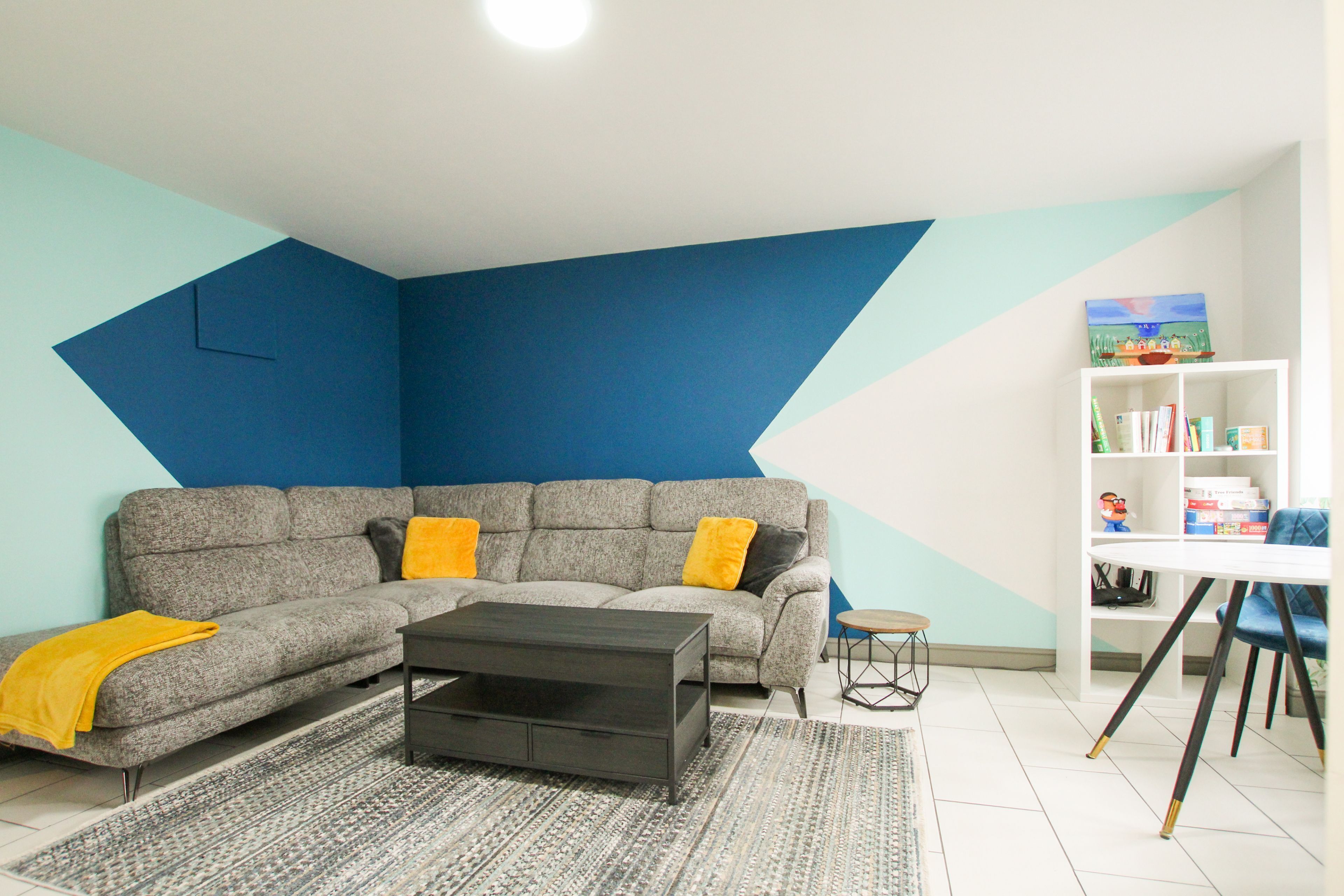 Ein Wohnzimmer mit Ecksofa, Couchtisch, Bücherregal und geometrischer Wandgestaltung.