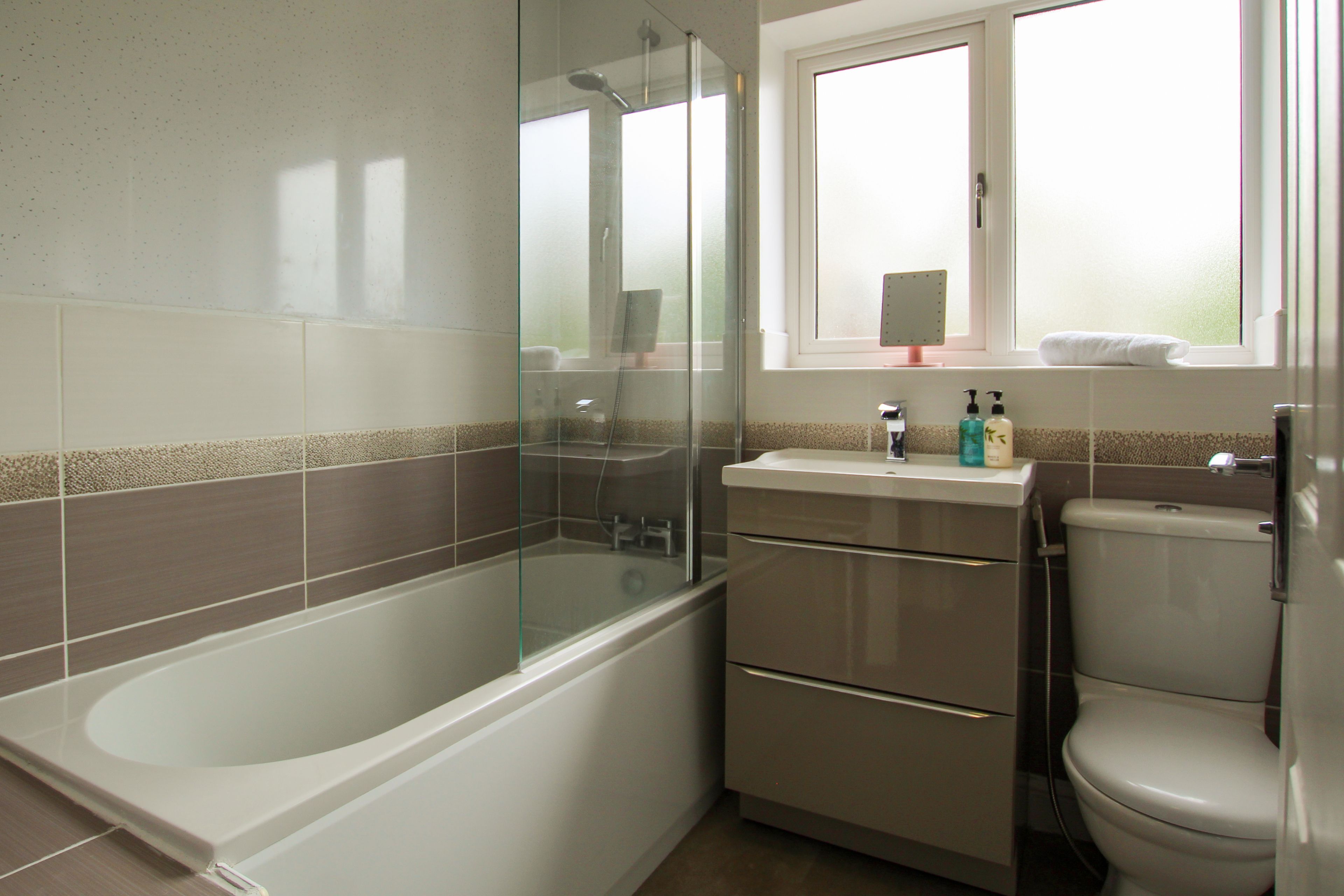 Une salle de bain moderne dotée d’une douche à parois de verre, d’une baignoire blanche, de toilettes et d’une fenêtre laissant entrer la lumière naturelle.