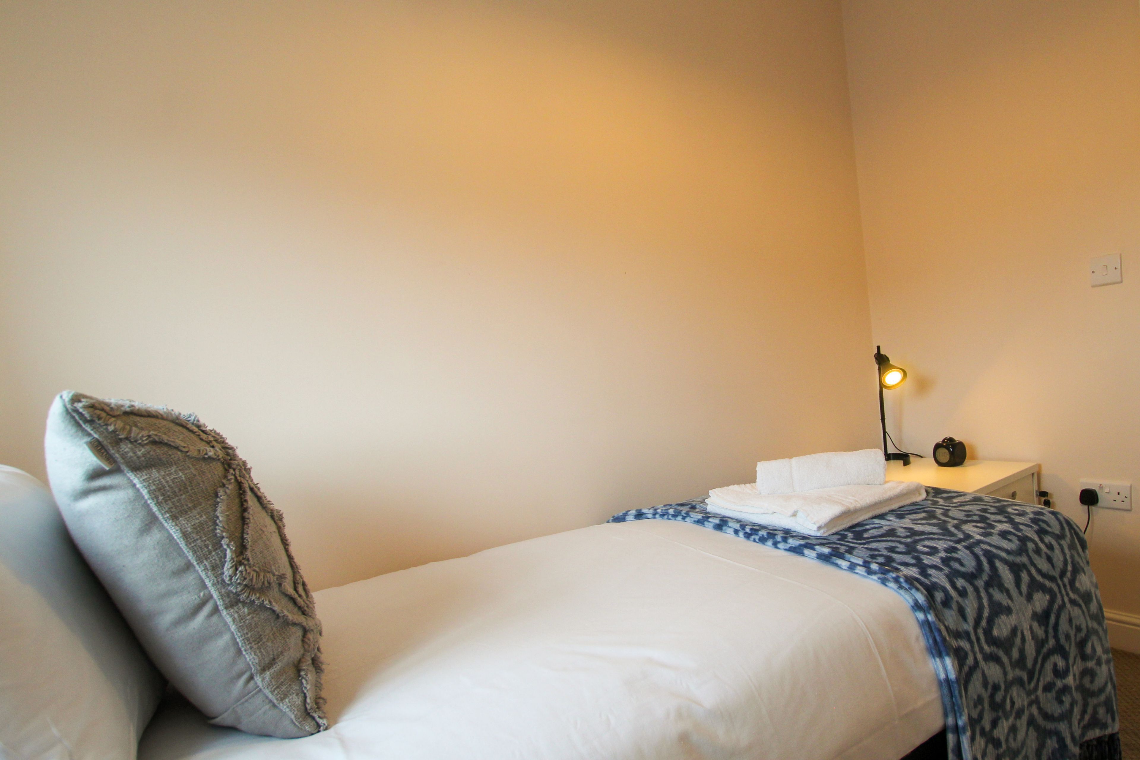 A single bed with a white comforter and a blue patterned blanket, a grey pillow, a bedside lamp, and a small table with a white towel and a black object.