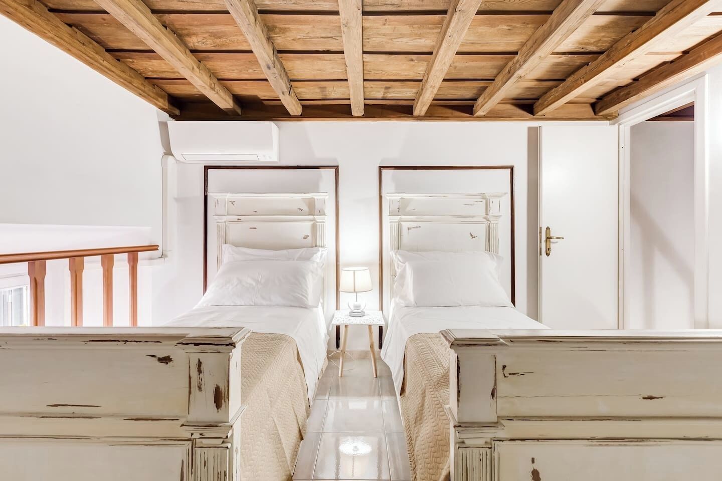 A room with two beds, a wooden ceiling, white walls, and a small table with a lamp.