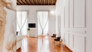 Ein geräumiges Wohnzimmer mit Holzboden, weißen Wänden und großen Fenstern mit durchsichtigen Vorhängen. Ein Kamin, ein Fernseher und zwei Sessel sind ebenfalls vorhanden.