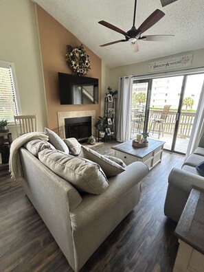 A living room with a sofa, a coffee table, a fireplace, a ceiling fan, and a view of the outdoors through a sliding glass door. - Beautiful 2-bedroom, 2 bath condo, located in Myrtle Beach, steps to the beach! (Myrtle Beach)