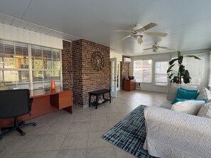 Un vaste espace de vie agrémenté d’un mur en briques, d’un ventilateur de plafond, d’un canapé avec des coussins turquoise, d’un bureau avec une chaise et d’une grande fenêtre avec des stores.