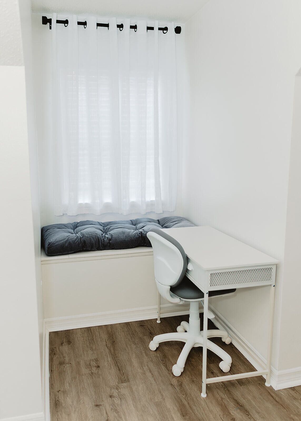 A small, white room with a wooden floor, a white desk with a chair, a window with sheer curtains, and a cushioned bench.