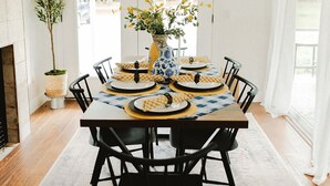A dining room with a large table set for six, surrounded by black chairs. A vase with yellow flowers is on the table, and a tall plant is in the corner.