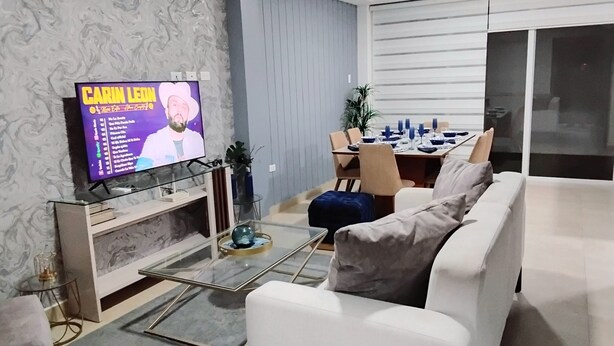Una sala de estar moderna con un televisor de pantalla plana, un sofá, una mesa de centro de vidrio y un comedor con una mesa lista para cuatro.
