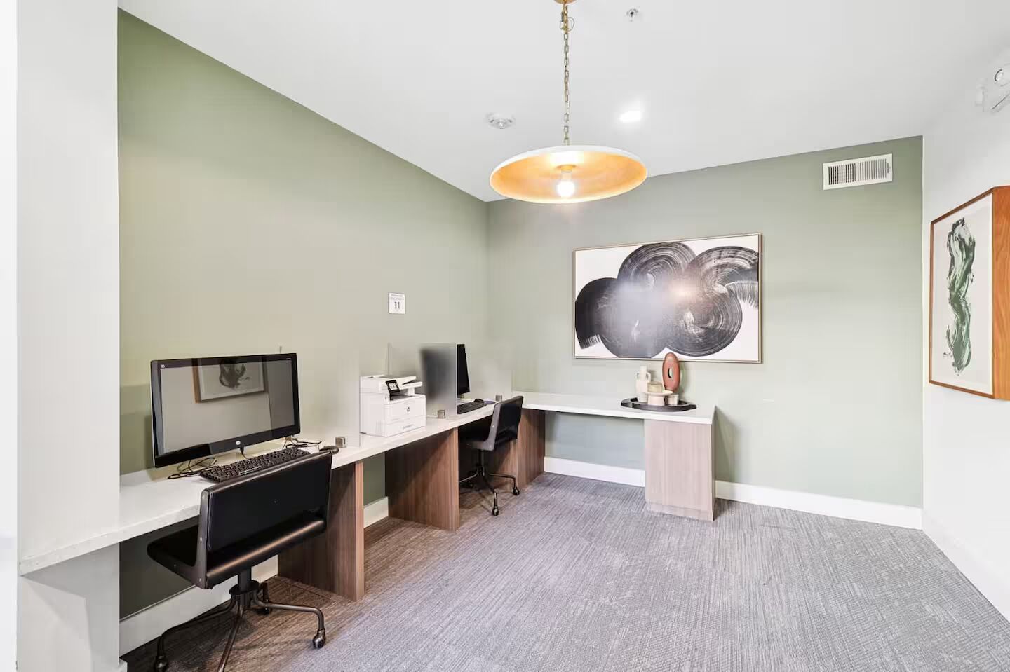 Un espace de bureau moderne comprenant un bureau, un ordinateur et une imprimante. Un grand tableau abstrait orne le mur, et un luminaire est fixé au plafond.