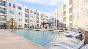 A swimming pool with lounge chairs and a waterfall feature, surrounded by multi-story buildings with balconies. - Lux 1B w Pool Near Toyota HQ (Frisco)