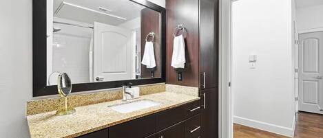 A bathroom with a granite countertop, a sink, a mirror, dark wooden cabinets, and a door leading to another room.