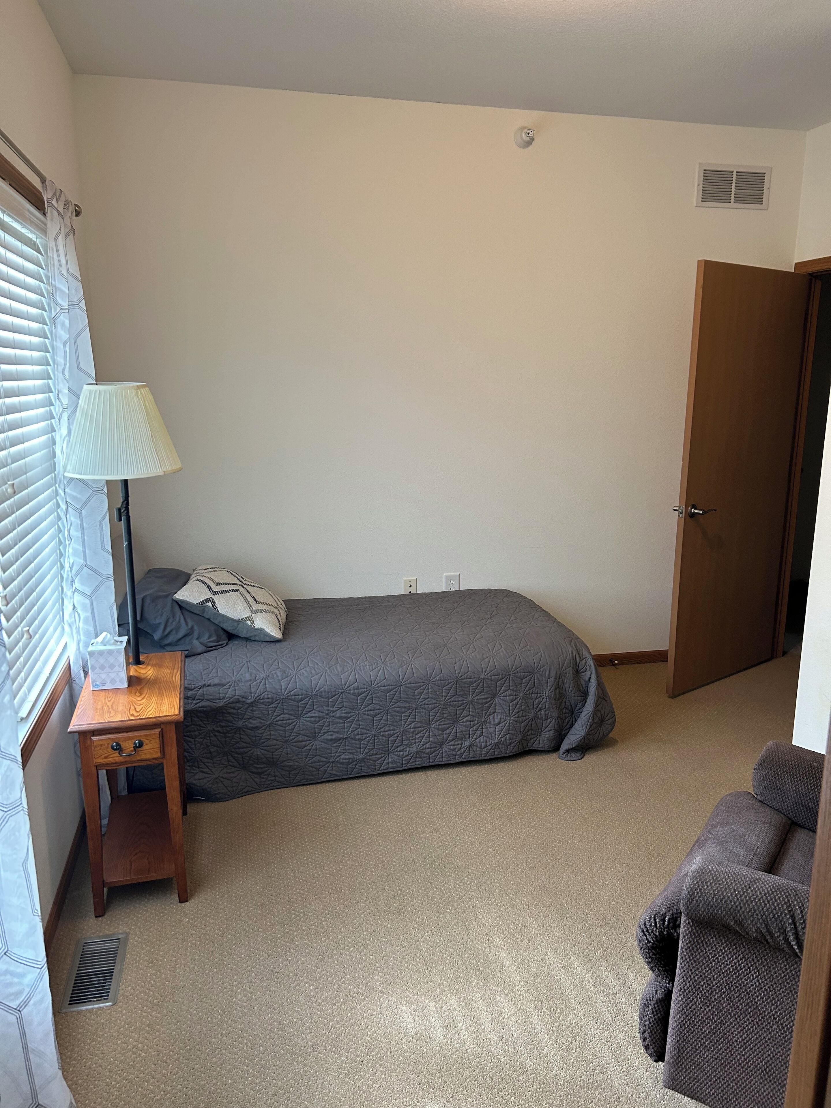 Une petite chambre à coucher, bien rangée, avec un lit simple, une table de chevet avec une lampe, une fenêtre avec des stores et une porte.
