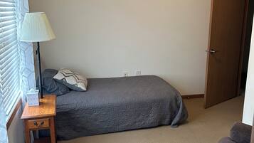 Une petite chambre à coucher, bien rangée, avec un lit simple, une table de chevet avec une lampe, une fenêtre avec des stores et une porte.