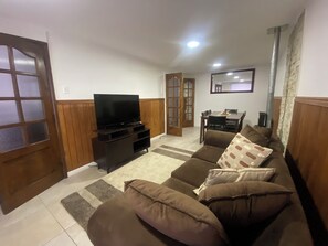 A living room with a flat-screen TV on a wooden stand, a brown sofa with patterned cushions, a dining table with chairs, and a mirror on the wall. - Spacious and comfortable house, excellent heating. (Punta Arenas)