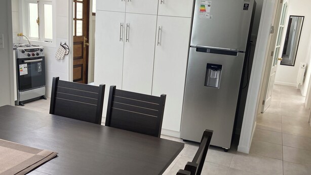 A modern kitchen with white cabinets, a stainless steel refrigerator, and a built-in oven. There is a dining table with chairs and a window above it. - Spacious and comfortable house, excellent heating. (Punta Arenas)