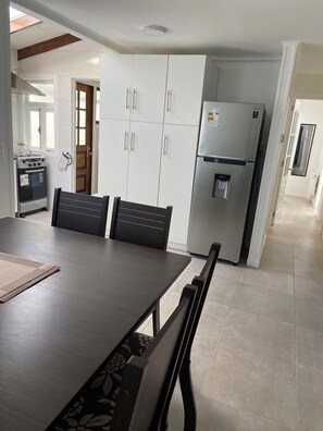 Een moderne keuken met witte kastjes, een roestvrijstalen koelkast en een inbouwoven. Er staat een eettafel met stoelen en erboven een raam.