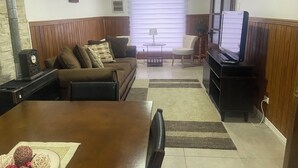 A living room with a sofa, a TV stand, a dining table with a placemat, and a window with blinds. - Spacious and comfortable house, excellent heating. (Punta Arenas)