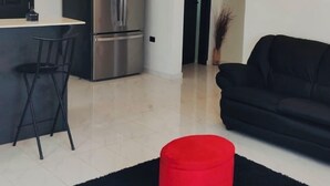 Una sala moderna con un sofá negro, un reposapiés rojo y una cocina con refrigerador de acero inoxidable.