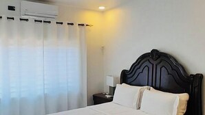 Un dormitorio con cama, ventilador de techo, equipo de aire acondicionado y mesita de noche con lámpara.