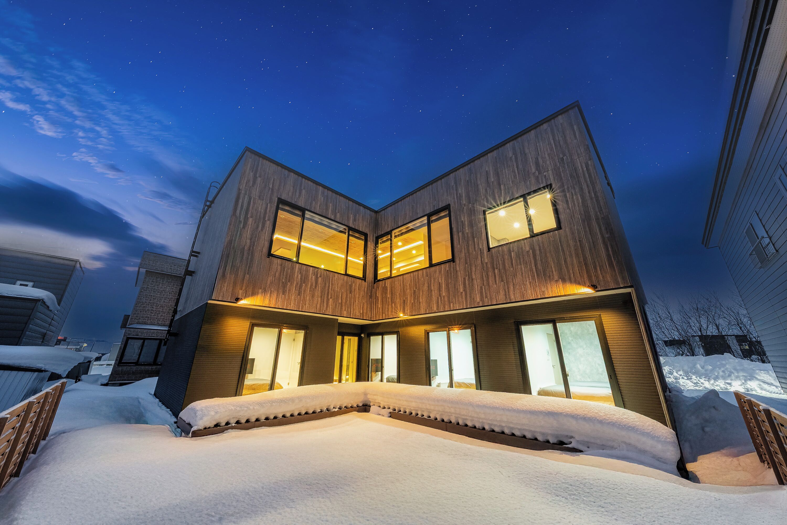 Una casa moderna de dos pisos con ventanas amplias y un camino de entrada cubierto de nieve.