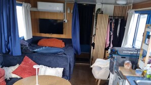 Ein kompaktes Wohnmobil-Interieur mit Bett, kleiner Küchenzeile, Tisch und an der Wand montiertem Fernseher.