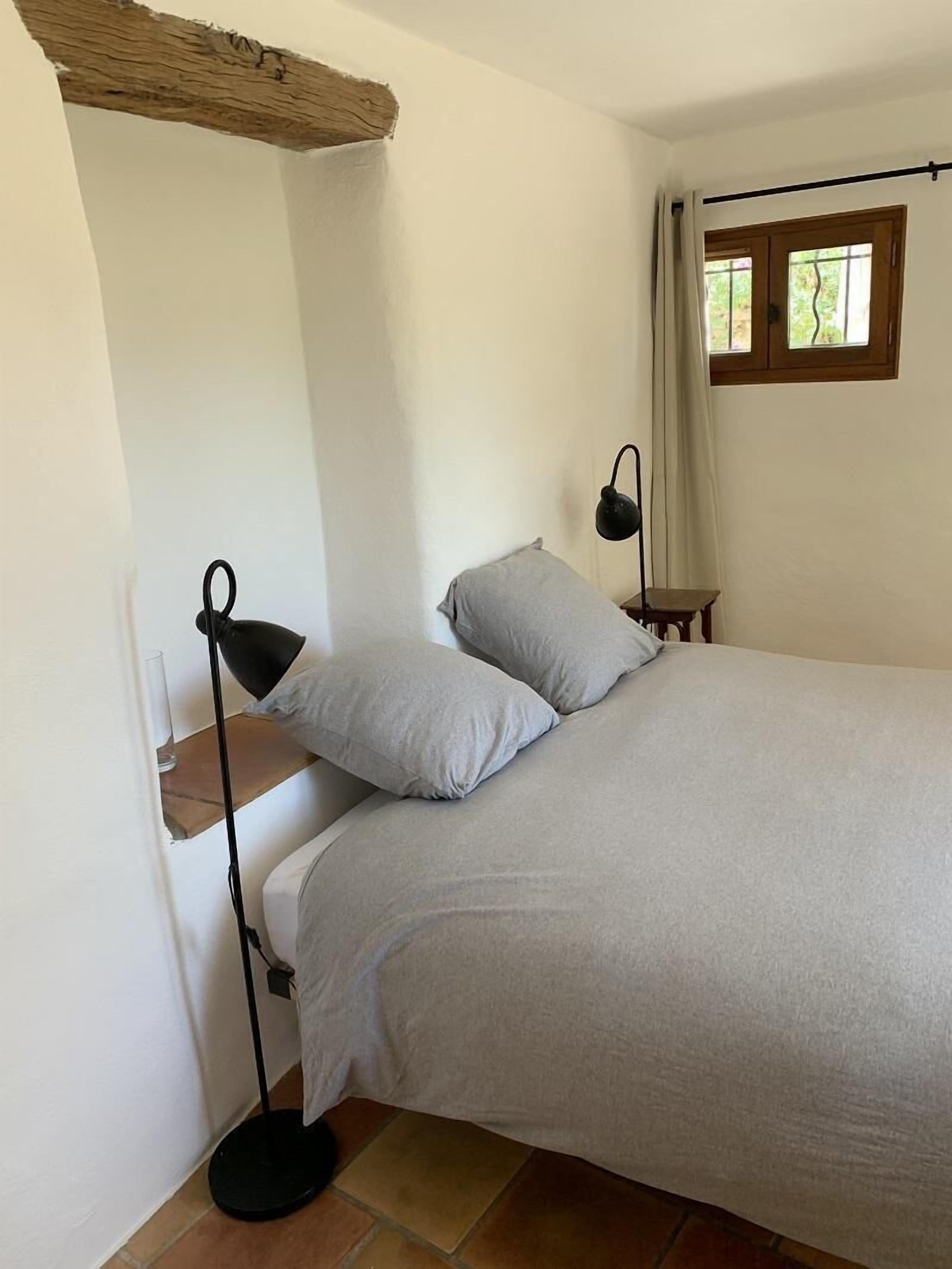 Een slaapkamer met een bed, kussens, een nachtkastje, een lamp en een raam met gordijnen.
