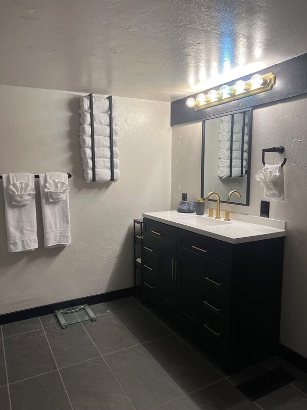 Un baño moderno con un lavamanos blanco, muebles oscuros y un espejo grande con luces arriba. Hay dos toalleros con toallas.