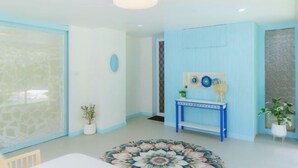 Une pièce dotée d’une grande fenêtre, d’une table ronde surmontée d’un tapis à motifs floraux, d’une console bleue et d’une plante en pot.